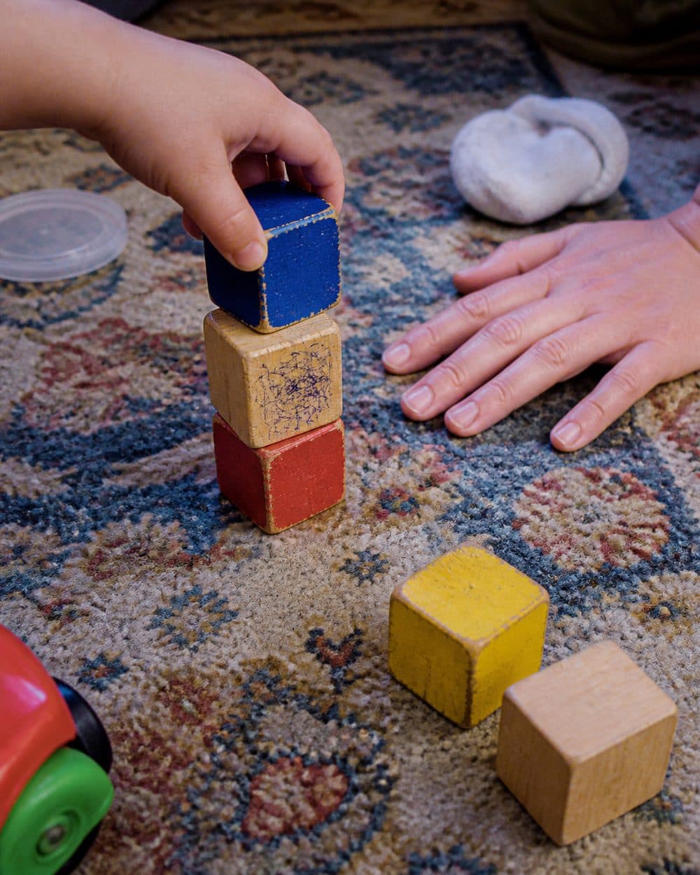 A child stacking a wooden block while an adult hand waits nearby