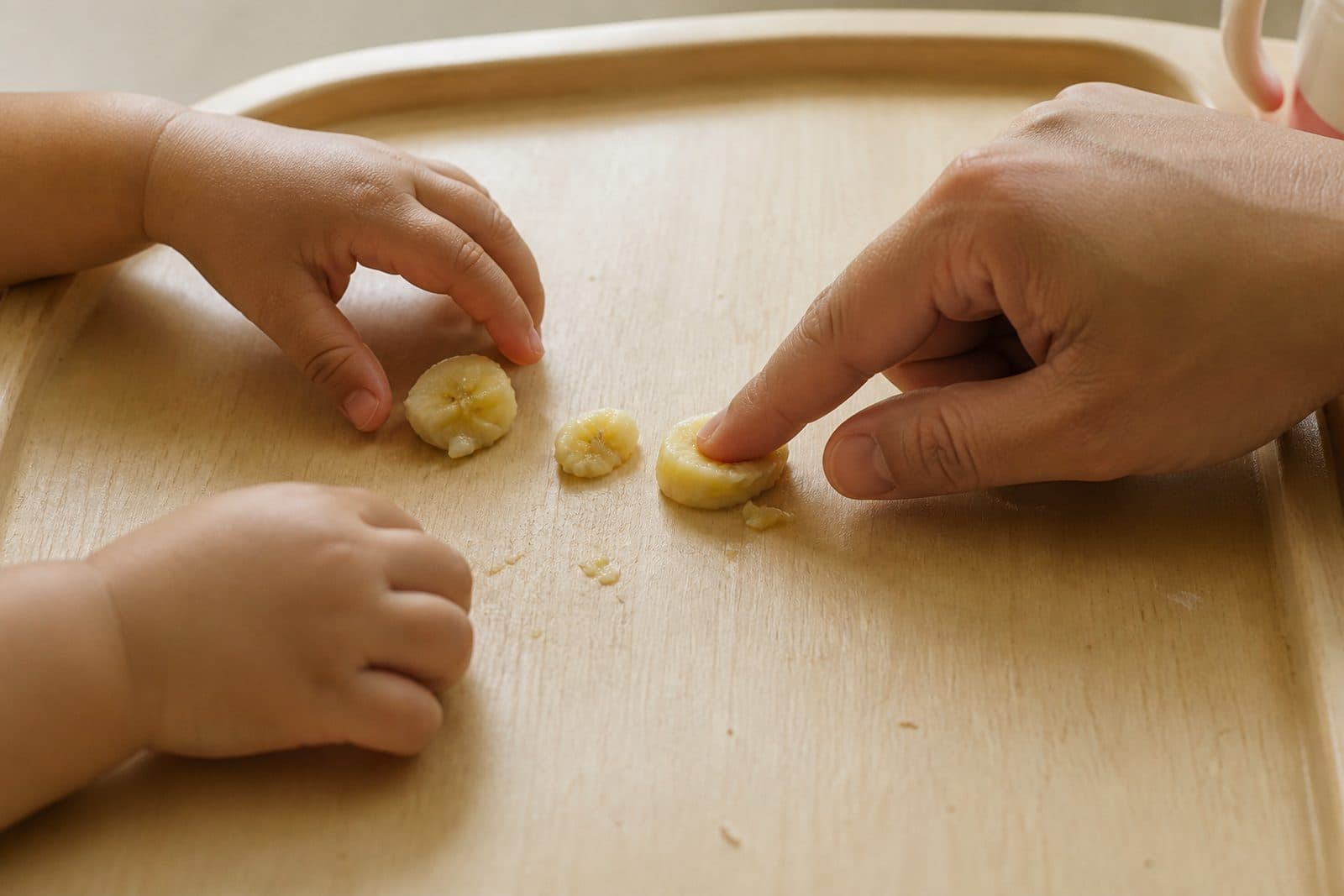 A toddler and a parent picking up pieces of banana on a highchair tray